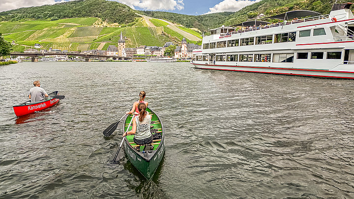 Das Foto zeigt zwei Kanus auf der Mosel bei Bernkastel-Kues. Rechts im Bild sieht man außerdem ein Personenschiff, das an einem Anleger hält. Die Kanufahrer haben einen tollen Blick auf die Weinberge von Bernkastel-Kues.