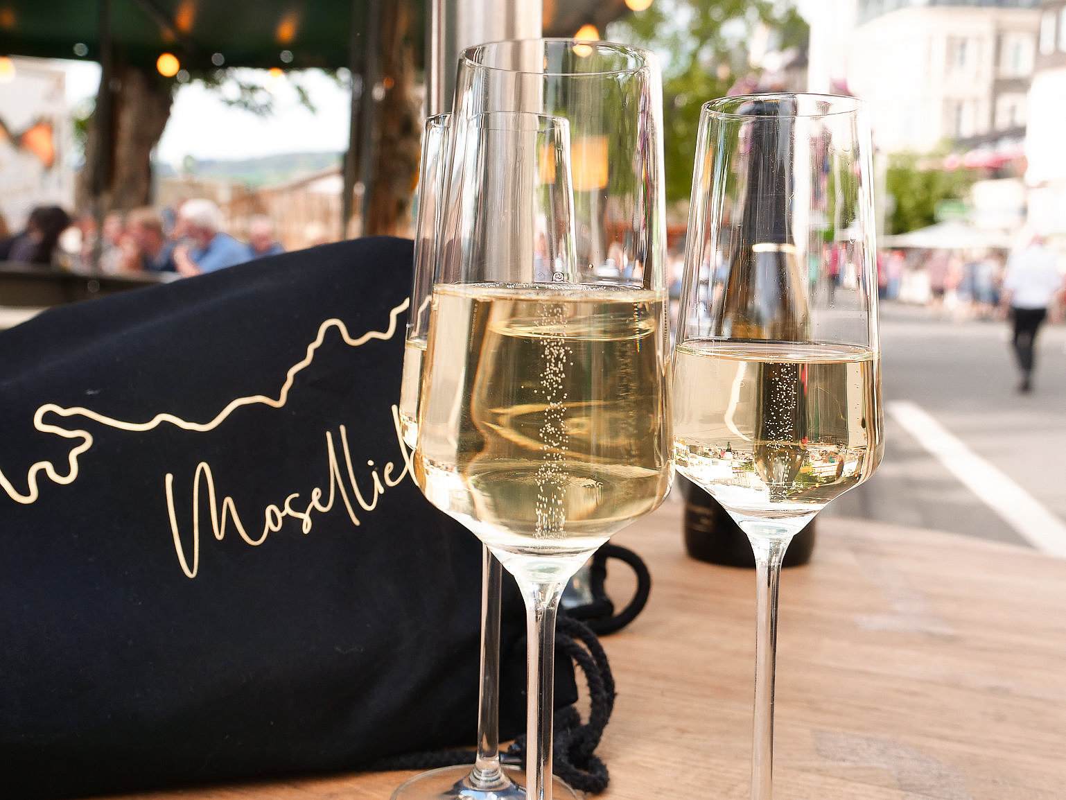 Moselliebe auf dem Weinfest der Mittelmosel in Bernkastel-Kues Das Foto zeigt drei SektglĂ€ser, die auf einem Holztisch stehen. Im Hintergrund ist die WeinstraĂe zu sehen.