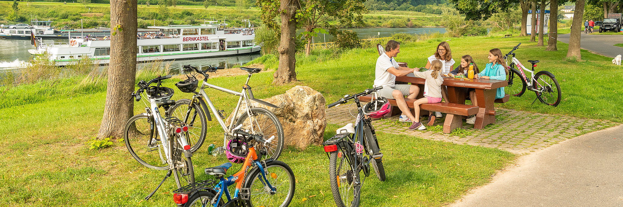 Ansicht auf eine Familie mit 3 Mädchen, die während ihrer Fahrradtour ein Picknick auf einer Bank mit Blick auf die Mosel und die Weinberge machen.