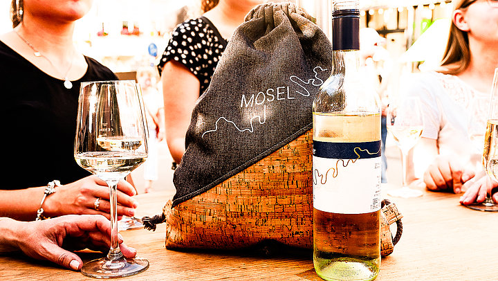Das Bild zeigt mehrere Personen, die an das Weinfest der Mittelmosel besuchen und an einem Tisch Wein genießen. Im Vordergrund steht ein Turnbeutel mit der Aufschrift Moselliebe und eine Flasche Wein.
