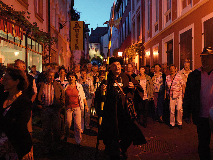 Das Foto zeigt den mit Mantel und Laterne verkleideten Nachtwächter, der eine Gruppe bei Dämmerung durch die Altstadt von Bernkastel führt.